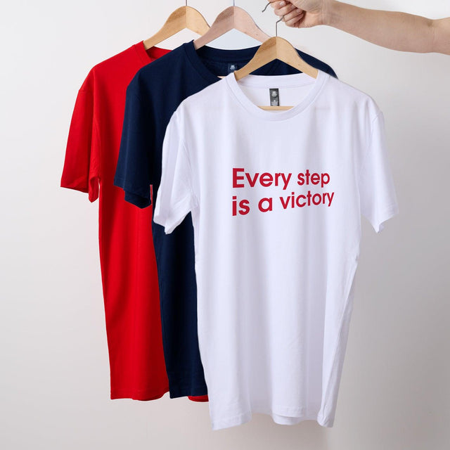 Three t-shirts hanging on wooden hangers, with the middle t-shirt being white and the others in red and blue. The white t-shirt has text 'Every step is a victory' on the front.