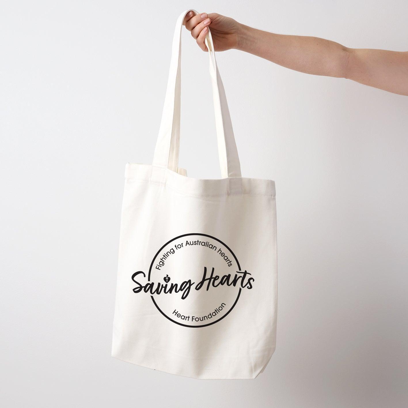 Heart Foundation cream canvas tote bag shown held in hand with Saving Hearts design in black print on the front.