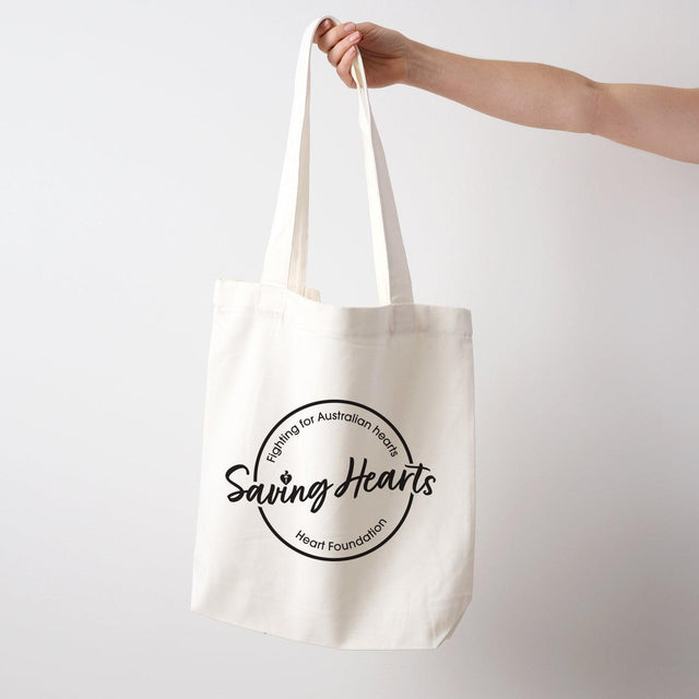 Heart Foundation cream canvas tote bag shown held in hand with Saving Hearts design in black print on the front.
