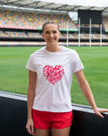 A smiling woman stands on a sports field wearing a white graphic T-shirt with a red heart-shaped slogan reading 'fight heart disease' and bright red athletic shorts, with stadium seating in the background.