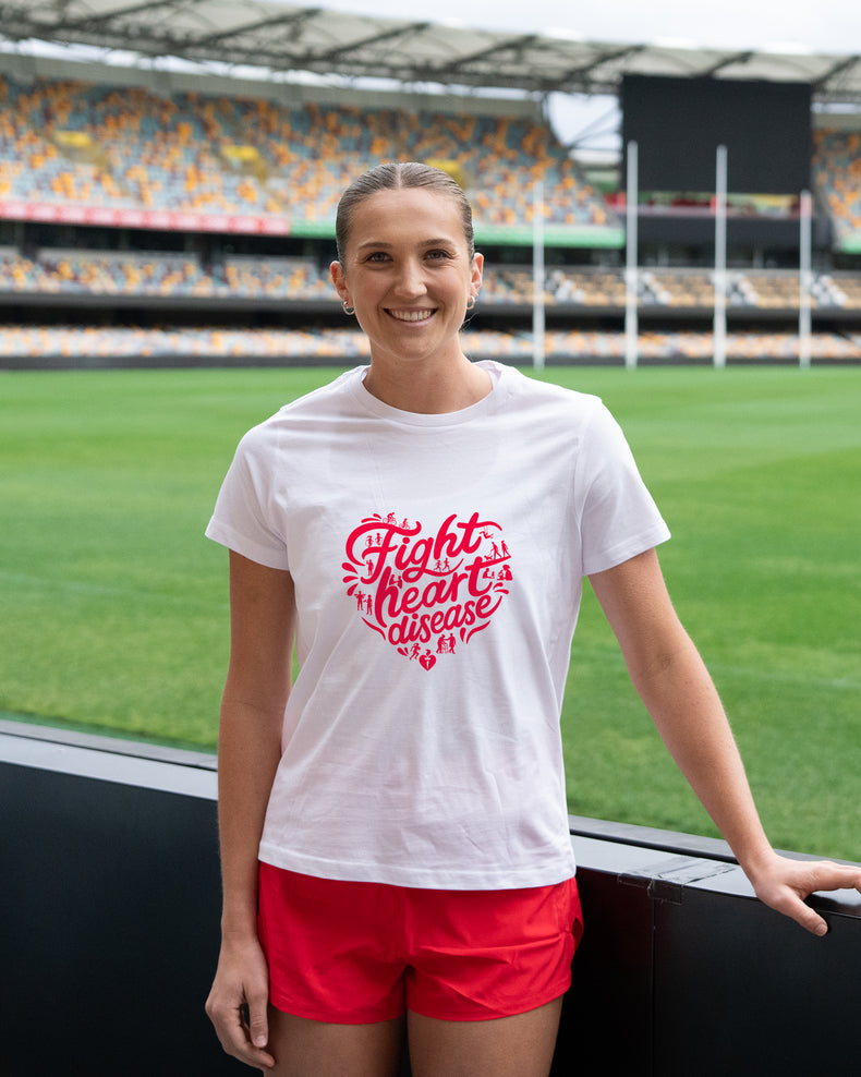 A smiling woman stands on a sports field wearing a white graphic T-shirt with a red heart-shaped slogan reading 'fight heart disease' and bright red athletic shorts, with stadium seating in the background.