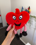 Red heart-shaped plush toy with a face held by a hand in an indoor setting.