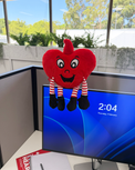 Red heart-shaped plush toy with legs and arms on a computer screen displaying a blue screen with time.