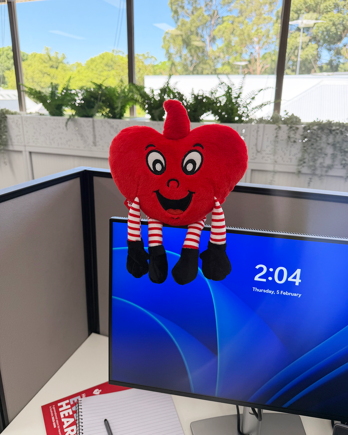 Red heart-shaped plush toy with legs and arms on a computer screen displaying a blue screen with time.