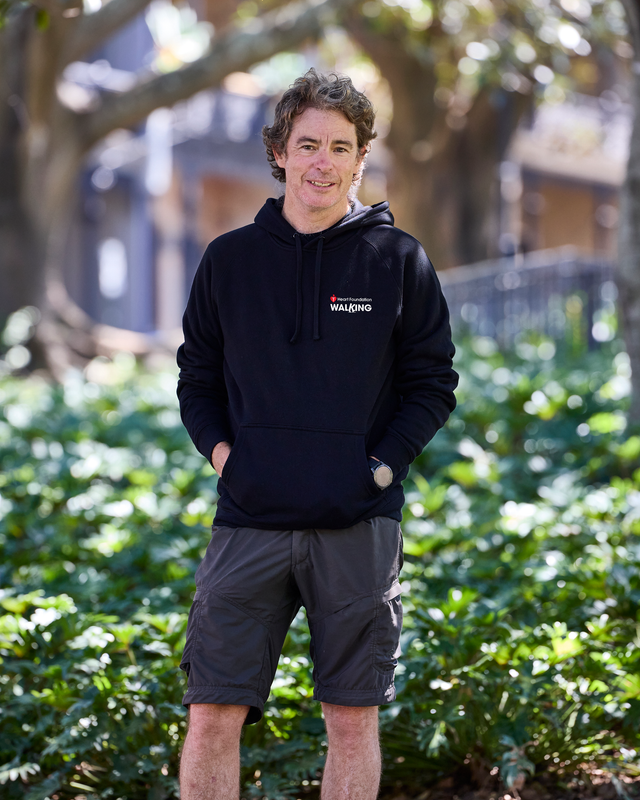 Man wearing a black hoodie with a logo standing outdoors among greenery
