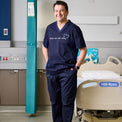 Nurse in navy scrubs standing in a hospital room with a Hill-Rom bed.