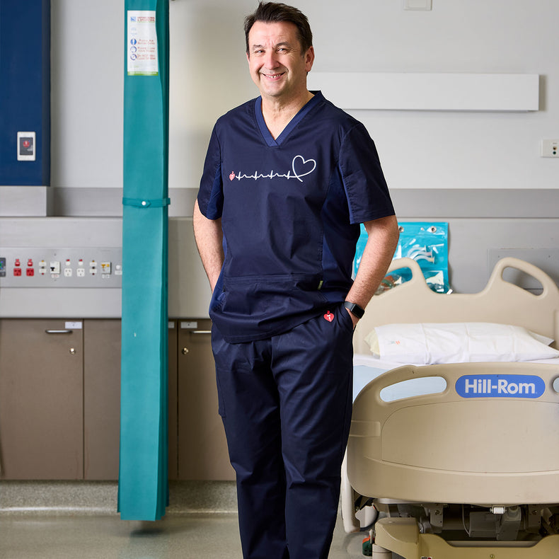 Nurse in navy scrubs standing in a hospital room with a Hill-Rom bed.