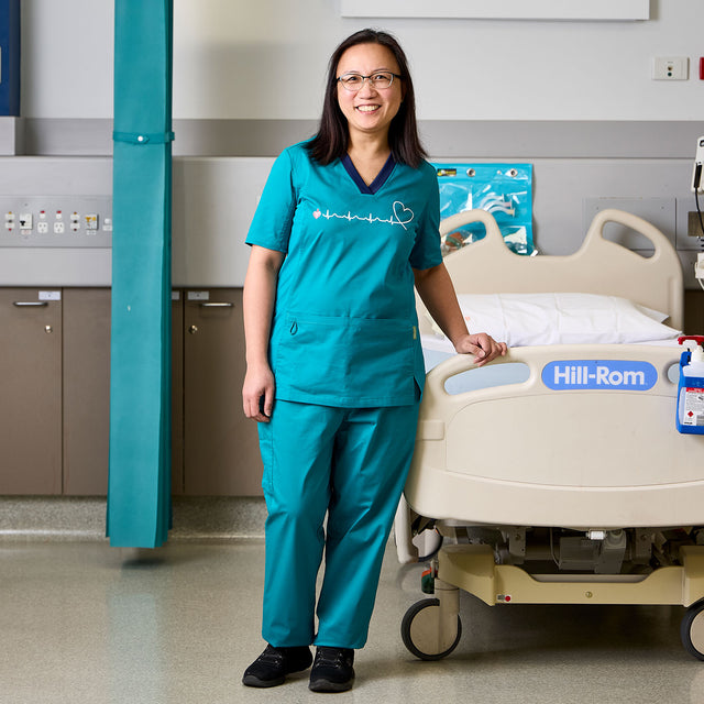 Nurse in teal scrubs standing next to a hospital bed with 'Hill-Rom' branding.