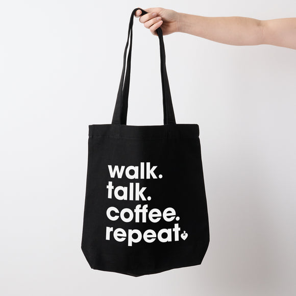 Heart Foundation black canvas tote bag shown held in hand with walk.talk.coffee.repeat. slogan in white print on the front.