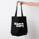 Heart Foundation black canvas tote bag held in hand with vintage look Heart hero text in white print. on the front