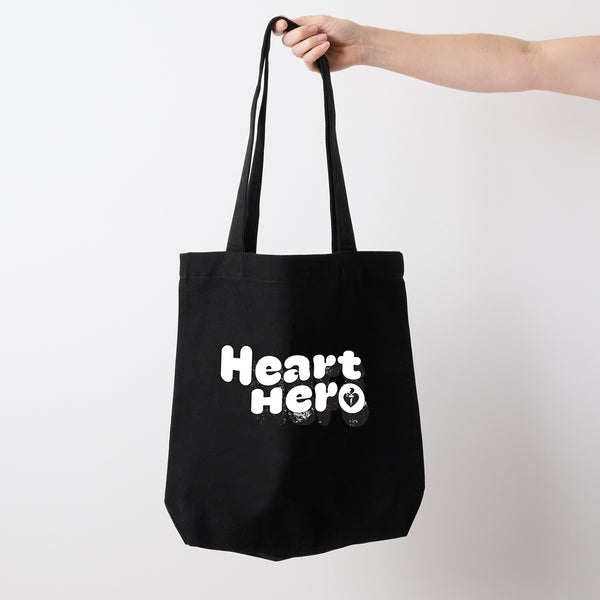 Heart Foundation black canvas tote bag held in hand with vintage look Heart hero text in white print on the front.