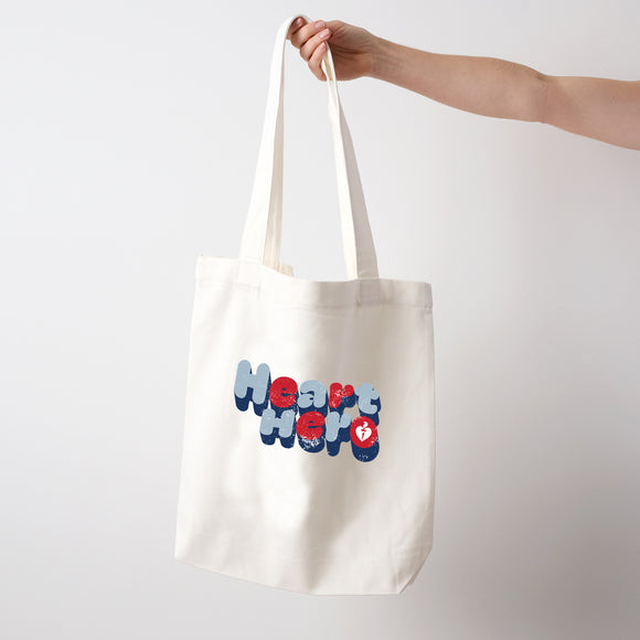 Heart Foundation cream canvas tote bag shown held in hand with vintage look Heart hero text in red and blue print. on the front