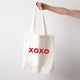 Heart Foundation white canvas tote bag shown held in hand  with XOXO design in red print on the front.